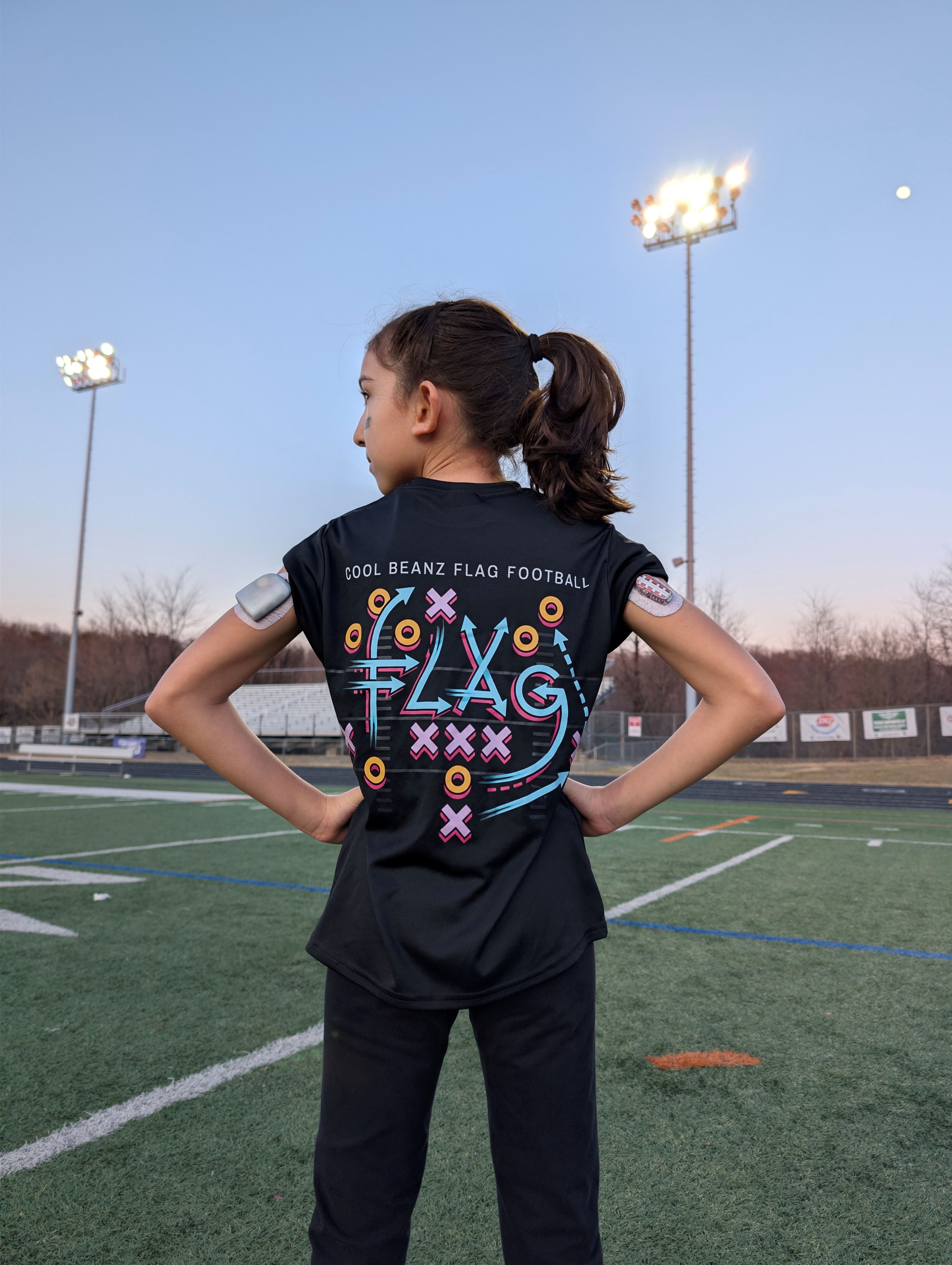 Young girl showing back of Cool Beanz Flag Football shirt with play design on field, youth flag football apparel and gear